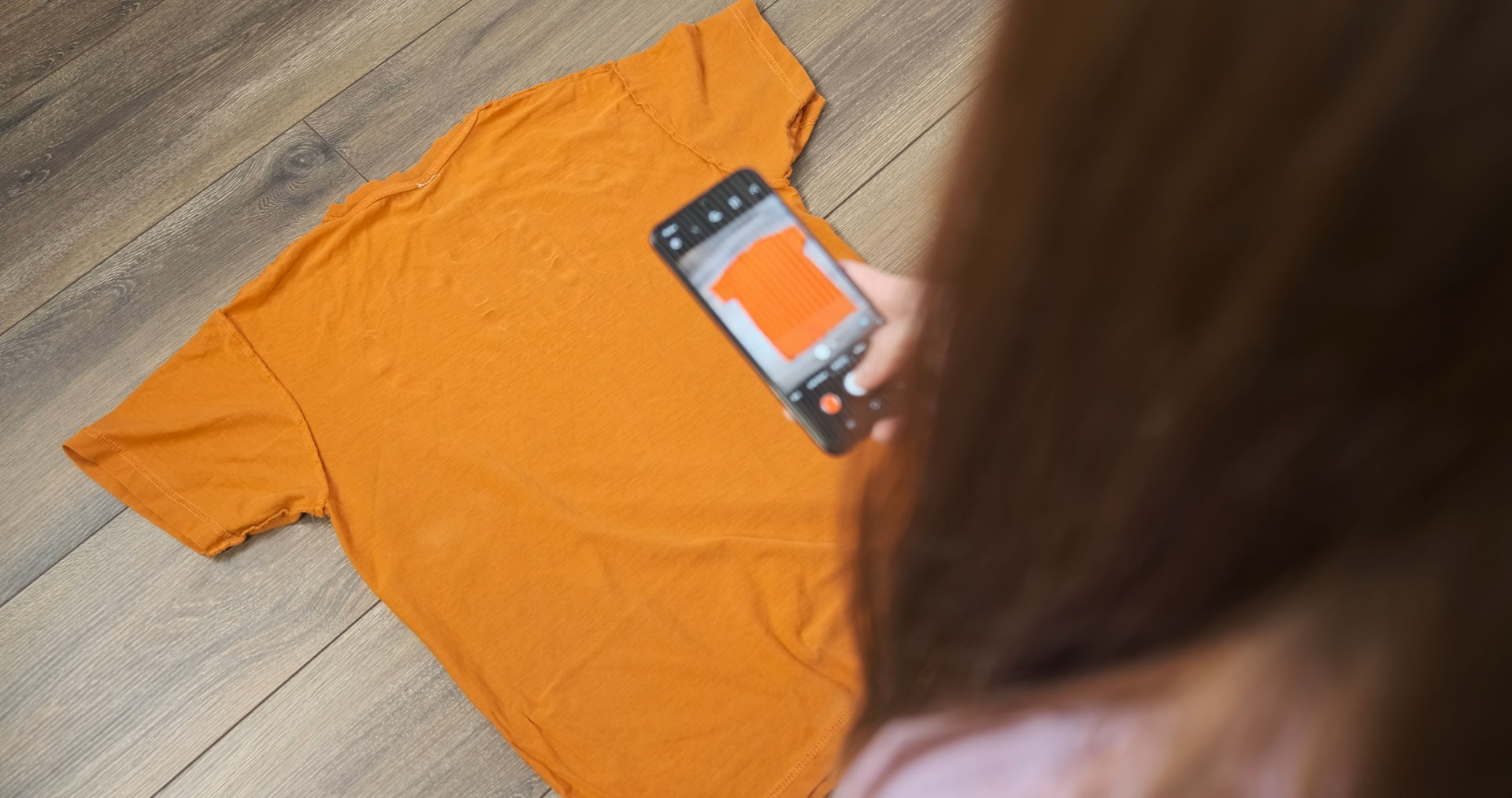 Resale of used items. Woman shoots t-shirt on the smartphone camera, spread out on the floor of the house. Back view.